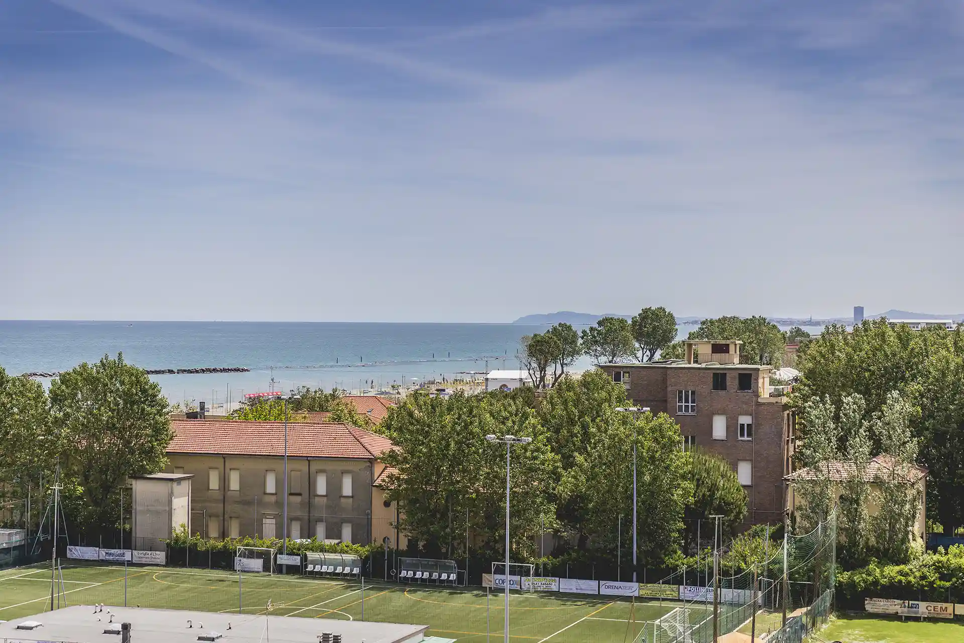 Blick auf eine Küstenlandschaft mit Gebäuden, Bäumen und einem Sportplatz im Vordergrund.
